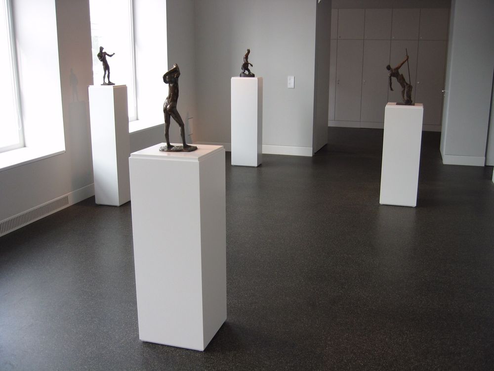 Weiße Ausstellungssockel mit Bronzeskulpturen in einem Galerieraum mit dunklem Boden
