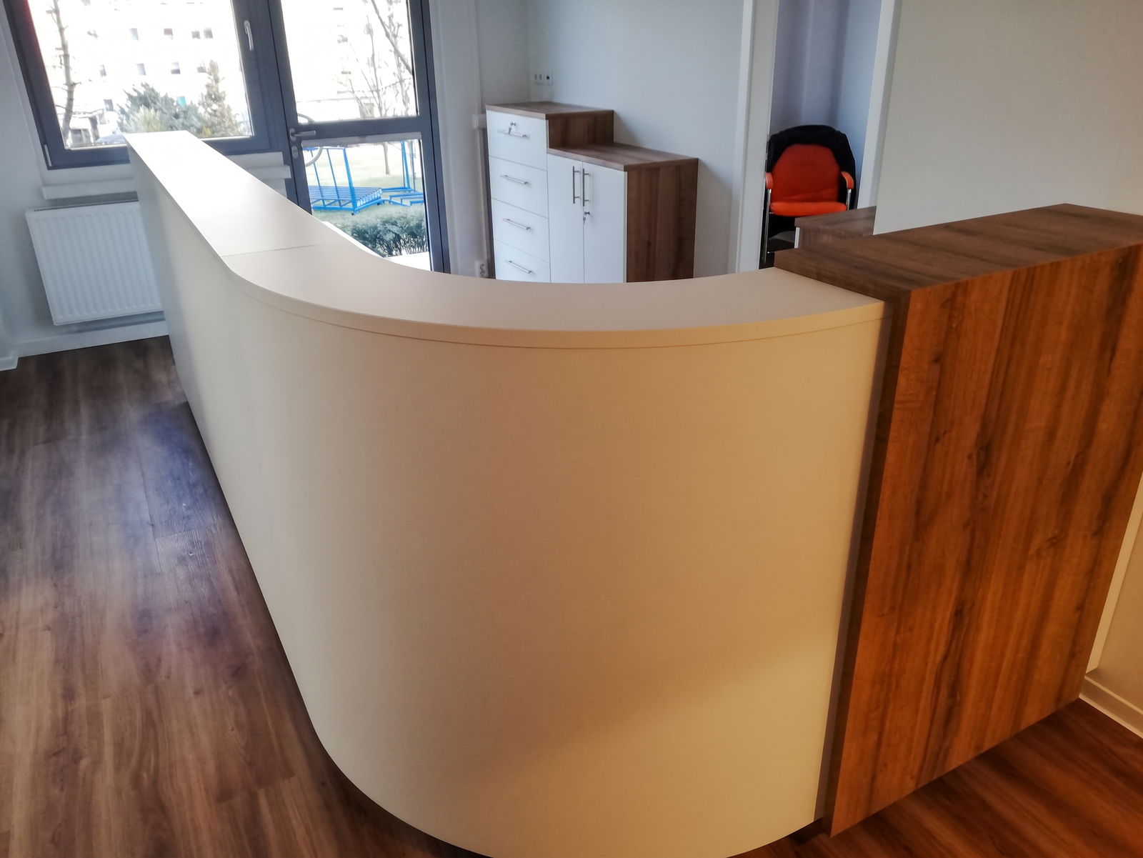 Geschwungene Empfangstheke in Creme mit Nussbaum-Stirnseite, dunkler Holzboden und Fensterfront