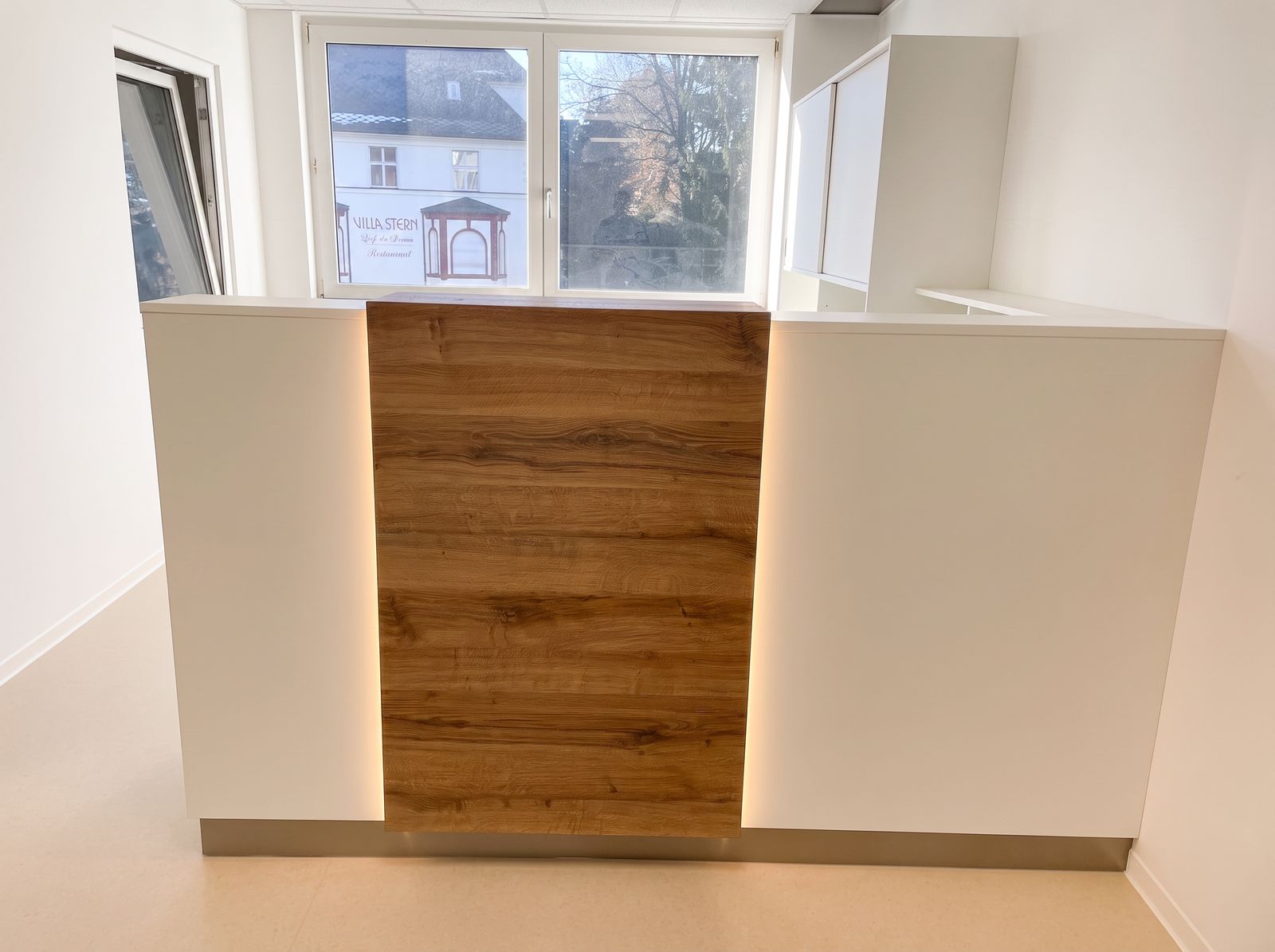 Weiße Empfangstheke mit Nussbaum-Frontpaneel und indirekter LED-Beleuchtung vor Fenster