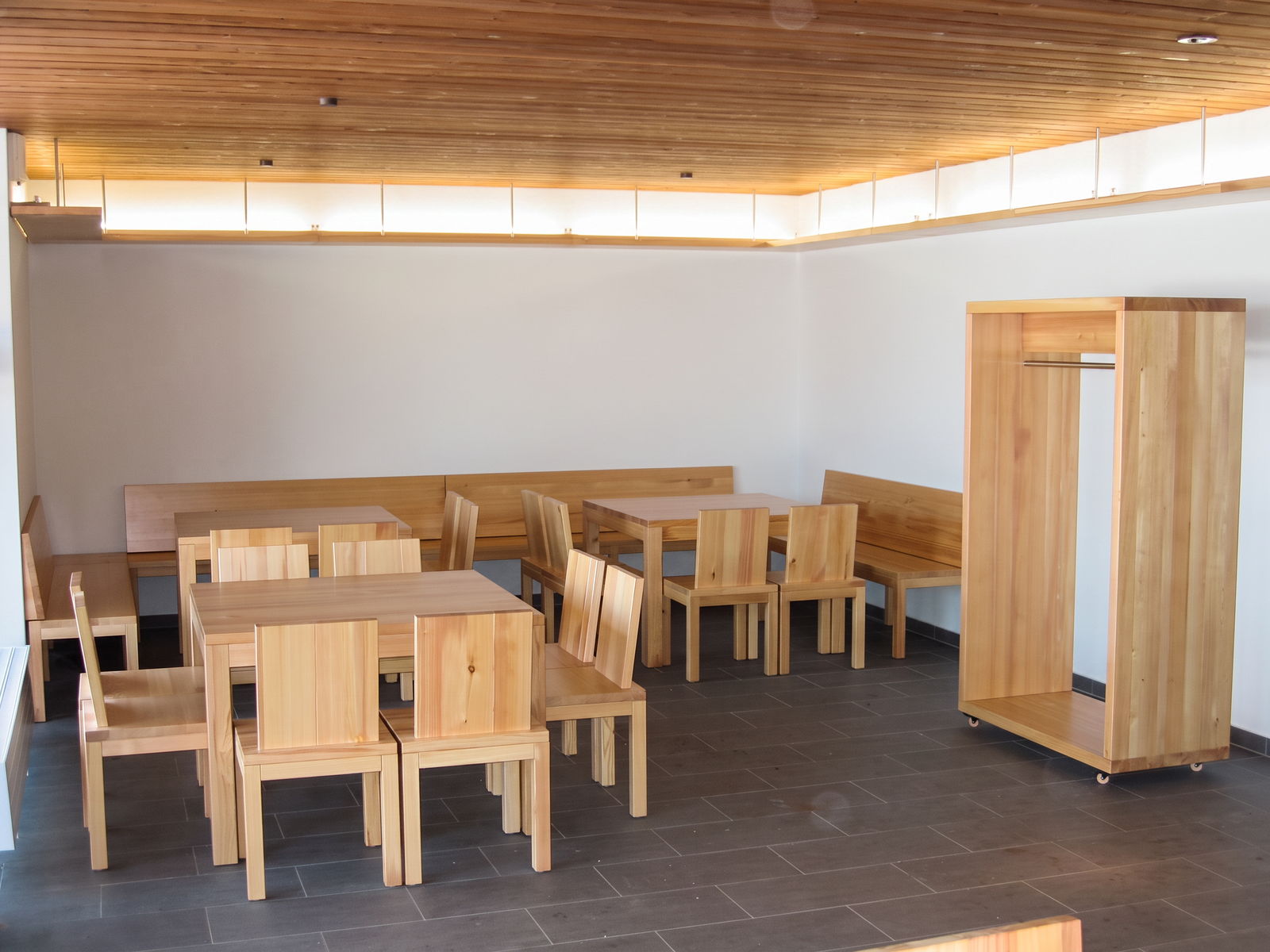 Speiseraum mit Tischen, Stühlen und Sitzbänken aus Massivholz, Holzdecke mit Lichtband und Garderobe auf Rollen