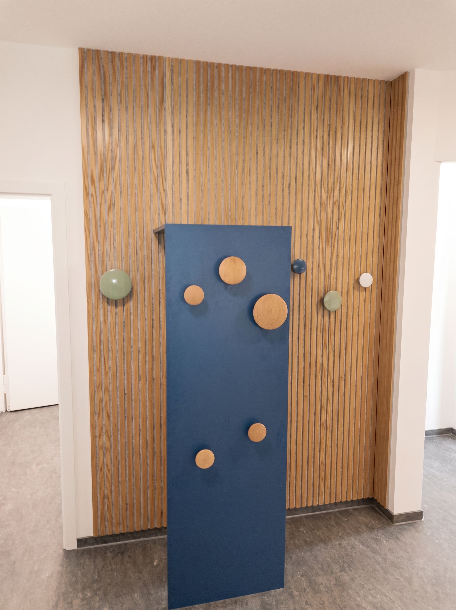 Garderobe mit Eichen-Akustiklamellenwand, blauem Paneel und runden Holzhaken in einer öffentlichen Einrichtung
