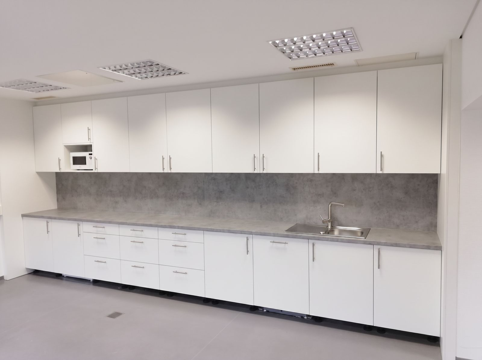 Weiße Einbauküche mit langer Arbeitsplatte, Rückwand in Betonoptik und Edelstahlspüle