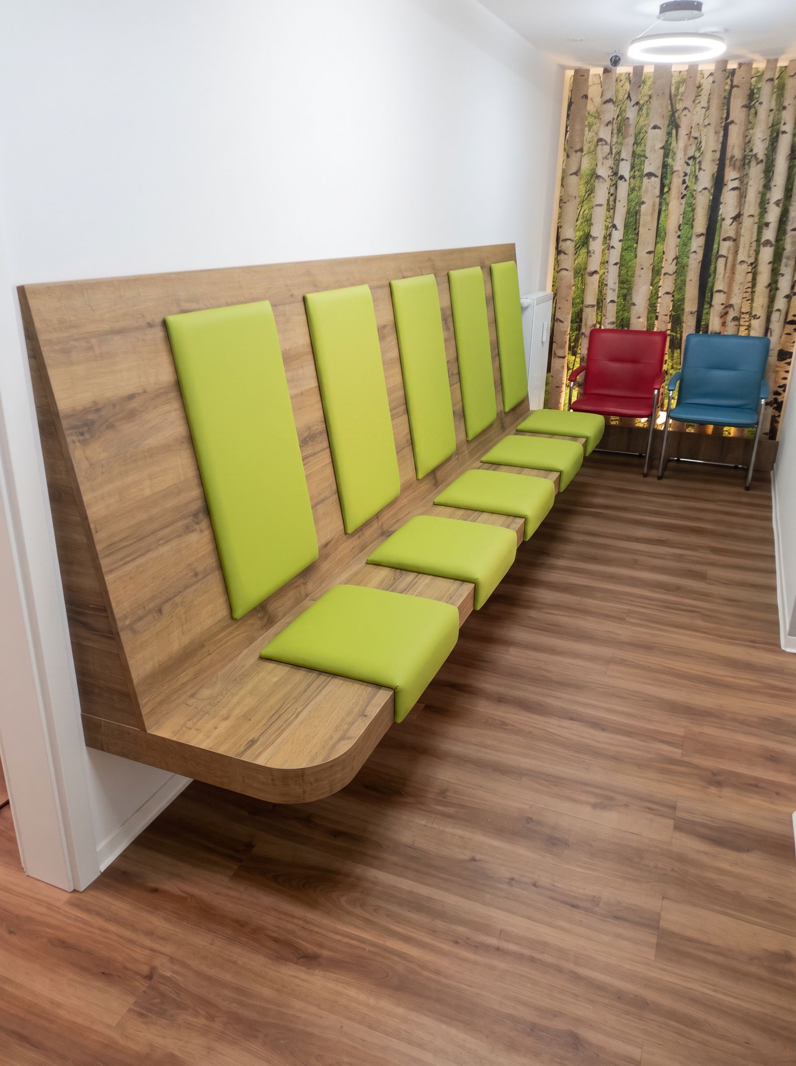 Wandhängende Wartebank aus Holz mit grünen Polstern, Wartebereich mit Birkenwald-Motivwand