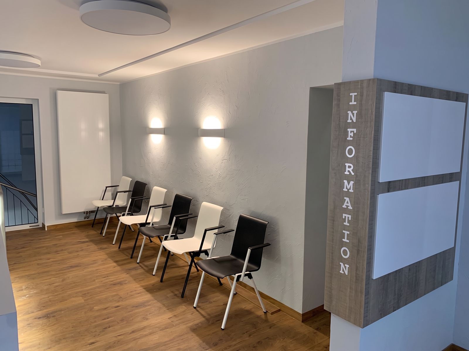 Wartebereich mit Stuhlreihe an der Wand, Wandleuchten und Informationssäule in Holzoptik