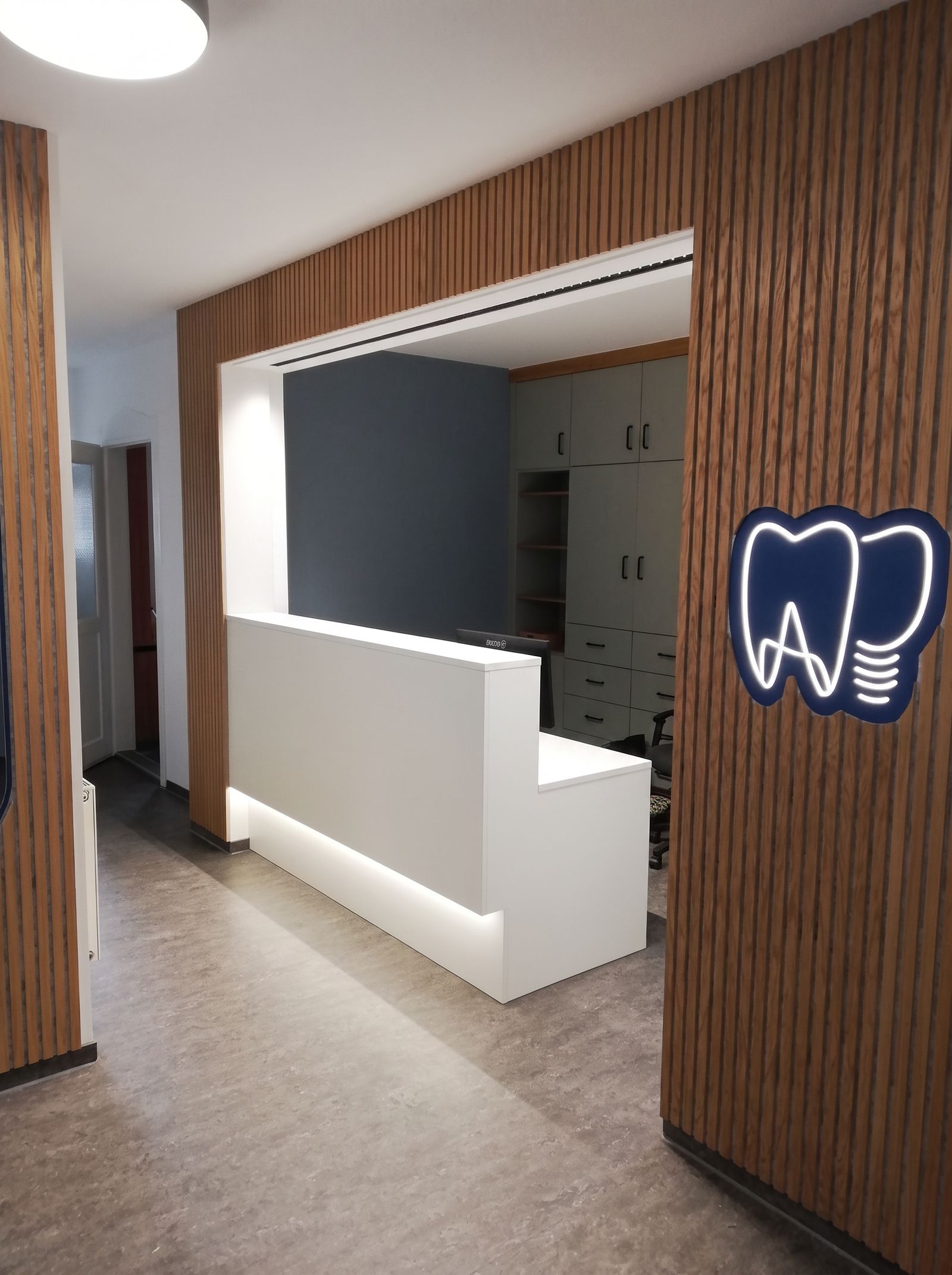 Zahnarzt-Empfang: weiße Theke mit LED-Sockel, Eichenlamellen-Wand und beleuchtetem Zahn-Logo